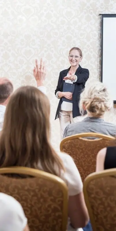 Intérieur d'une salle : lors d'une conférence, les personnes assises lèvent la main pour poser une question à la personne qui se trouve devant elles.